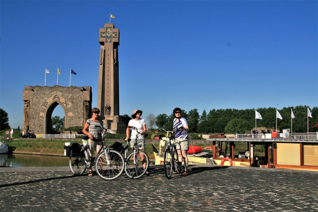 Die Radfahrer stehen neben dem Boot auf dem Kai. Im Hintergrund links sind das Friedenstor und der IJzer-Turm von Diksmuide zu sehen.